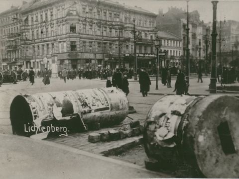 Auf dem Bild sind umgestürzte Litfaßsäulen zu sehen, welche als Barrikaden genutzt wurden. Im Hintergrund ist eine sich verteilende Menschenmege zu sehen, welche von der Kamera wegläuft.