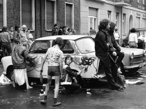 Die Fotografie von Marion Klemp zeigt eine Straßenszene in Ost-Berlin aus den 80er-Jahren. Es sind mehrere Fahrzeuge der Marke Trabant zu sehen, wovon einer von Kindern mit Farbe beschmiert wird. Zwei Erwachsene sind an das Fahrzeug angelehnt und schauen in eine andere Richtung.