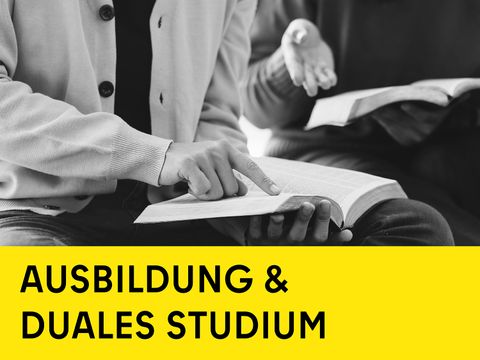 2 Personen, eine liest und fährt mit dem Finger über Buchseite, gelber Balken unten mit schwarzer Schrift Ausbildung & Duales Studium