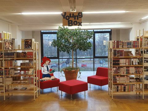 Jugendbereich der Mark-Twain-Bibliothek - Bücherregale, rote Hocker und Sessel und eine gezeichnete Mächenfigur, die mit untergeschlagenen Beinen auf einem der Sessel sitzt und in einem Buch liest