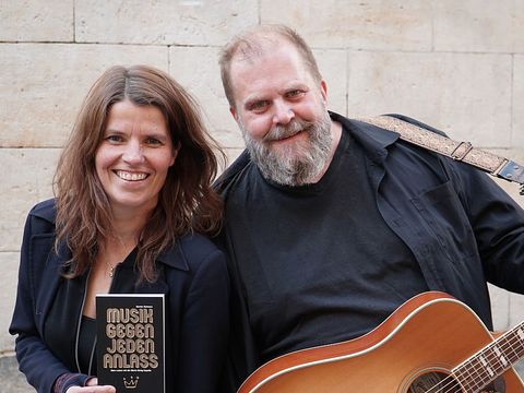 'Maria König' und 'Knut Schfleck' lachend vor einer Steinwand. Maria hält das Buch 'Musik gegen jeden Anlaß' in den Händen, Knut hat eine Akustikgitarre umghängt qua