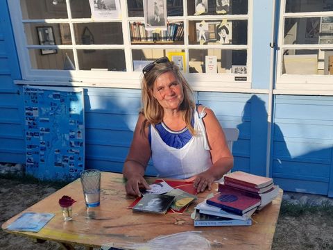 Das Bild zeigt die Autorin Ute Fritsch vor ihrem Haus auf Hiddensee. Sie sitzt umringt von ihren Kunstwerken und Büchern.
