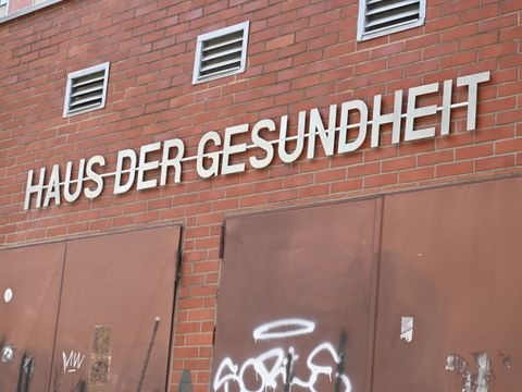 Außenansicht eines Gebäudes mit einer Klinkerfassade aus roten Ziegeln. An der Wand über braunen, mit Graffiti beschmierten Toren ist der Schriftzug ‚HAUS DER GESUNDHEIT‘ in großen, weißen Großbuchstaben angebracht.