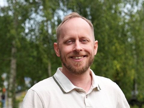Porträtfoto von Michael Kusserow, Leiter der Webredaktion des Bezirksamtes Marzahn-Hellersdorf. Er blickt lächelnd in die Kamera und trägt ein beiges Poloshirt mit dunklen Akzenten am Kragen und den Ärmeln. Der Hintergrund mit grünen Bäumen ist unscharf.