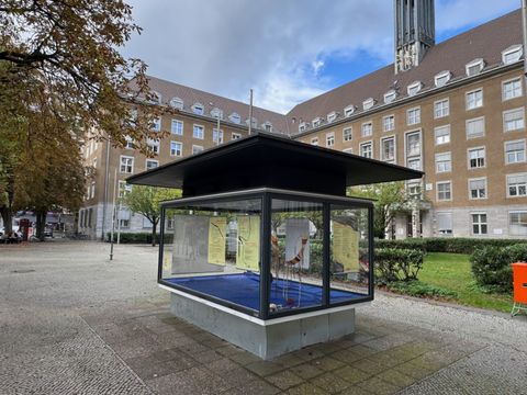 Die gläserne Ausstellungsvitrine auf dem Mathilde-Jacob-Platz, dahinter das Rathaus Tiergarten mit seinem Uhrenturm