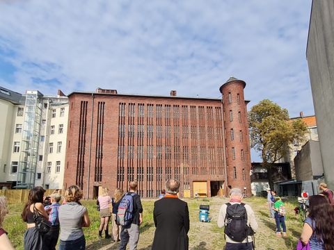 Menschen stehen auf einem Feld vor einem alten Backsteingebäude in der Sonne und schauen auf das alte Gebäude