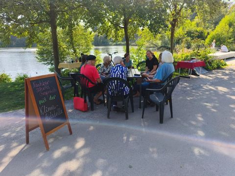 Das Foto zeigt eine Gruppe von 7 Personen, die sich an einem Tisch sitzend, unterhalten. Im Hintergrund ist ein See und Bäume zusehen