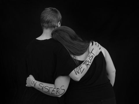 Zwei Frauen in Schwarz Weiß vor einem Schwarzen Hintergrund. Sie nehmen sich seitlich in den Arm. Die eine Frau hat ihren Kopf auf die Schulter der anderen gelegt. Auf den Armen der Frauen steht geschrieben "In Sisters we Trust" in Großbuchstaben.