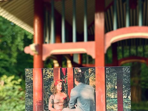 Botanischer Garten – Frau Holles Garten: Hand hält Foto einer Filmszene mit zwei Personen im roten Pavillon in die Kulisse des Drehorts