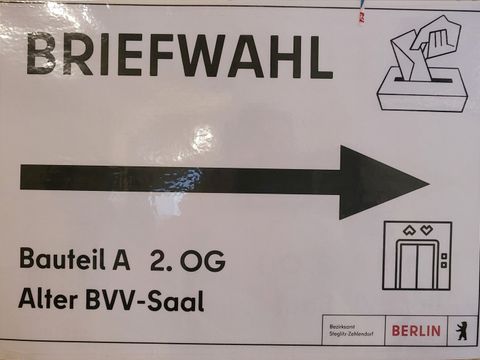 Wegweiser zum Briefwahl-Büro im Rathaus Zehlendorf, Bauteil A, 2. OG, Alter BVV-Saal mit dickem schwarzen Pfeil, Piktogramm: Urne mit Schlitz und Briefumschlag, Piktogramm: Fahrstuhl