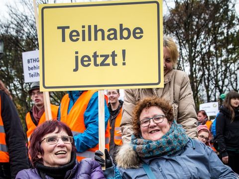 Bild von der Demonstration: Nicht mein Gesetz, zwei Frauen sitzen eng nebeneinander, mit dicken Jacken (eine lila, die andere petrol), Brillen, kurzen dunklen Haaren. Sie halten ein Schild auf dem steht auf gelben Hintergrund in schwarzer Schrift Teilhabe jetzt!