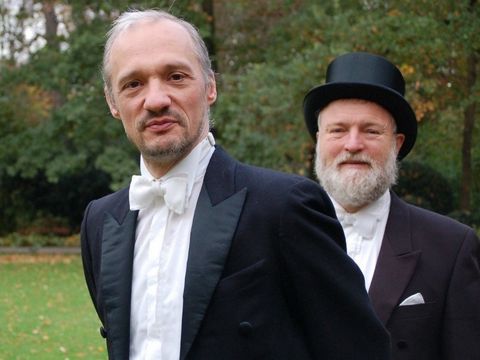 Zwei Herren in Frack, einer auch mit Zylinder, stehen im Park und schauen in die Kamera.