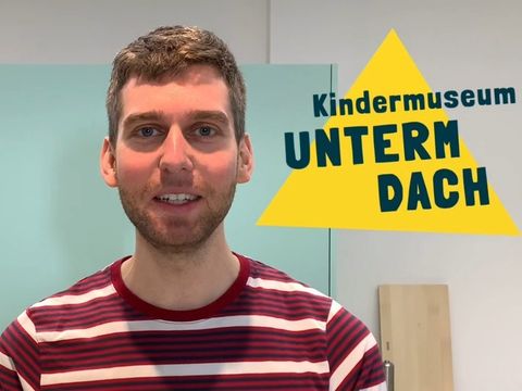 Ein Mann schaut in die Kamera. Neben ihm ist ein gelbes Dreicks-Logo mit dem Schriftzug "Kindermuseum unterm Dach" abgebildet.