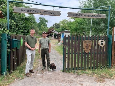 Zwei Männer und ein Hund stehen vor dem Eingang eines Grundstücks. Über dem Eingang steht auf zwei Schildern "Naturschutz Station Grünes Klassenzimmer".