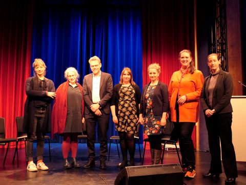 Sechs Frauen und ein Mann stehen nebeneinander für ein Gruppenfoto vor roten und blauen Samtvorhängen.