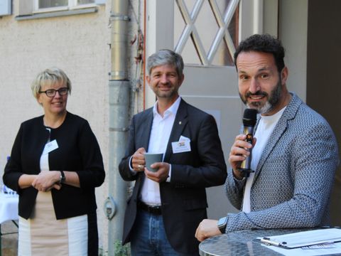 Zwei Männer und eine Frau stehen vor einer Hauswand und schauen in die Kamera. Ein Mann spricht in ein Mikrofon