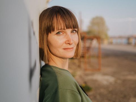 Eine Frau lehnt mit dem Rücken an eine Wand. Sie schaut über ihre Schulter in die Kamera und lächelt.