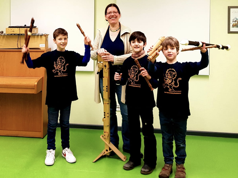 Eine Frau steht zusammen mit drei Kindern in einem Raum. Die Kinder halten Blackflöten in die Höhe.