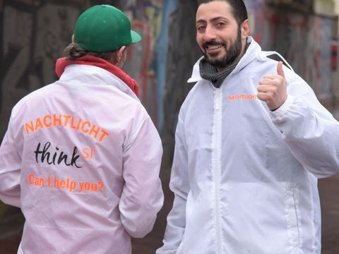 Zwei Personen stehen nebeneinander auf einem Gehweg und tragen weiße Jacken. Die linke Person ist mit dem Rücken zur Kamera gedreht und auf der Jacke steht "Nachtlicht think SI³ Can I help you?". Die rechte Peron lächelt in die Kamera und zeigt den Daumen nach oben.