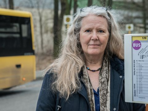 Anne Frau steht neben einer Bushaltestelle. Im Hintergrund steht ein Bus.