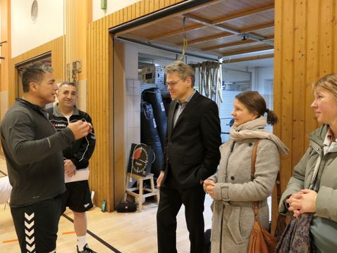 Zwei Männer unterhalten sich in einer Turnhalle. Zwei Frauen und ein Mann hören zu.
