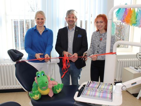 Zwei Frauen und ein Mann schneiden ein rotes Band vor einem Zahnarztstuhl durch.