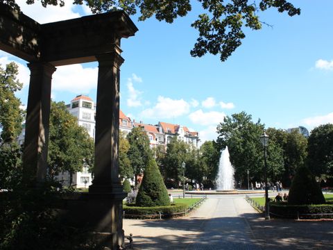 Zu sehen ist eine Ansicht des Viktoria-Luise-Platzes von den Kolonnaden aus. In der Mitte der Springbrunnen mit Fontäne
