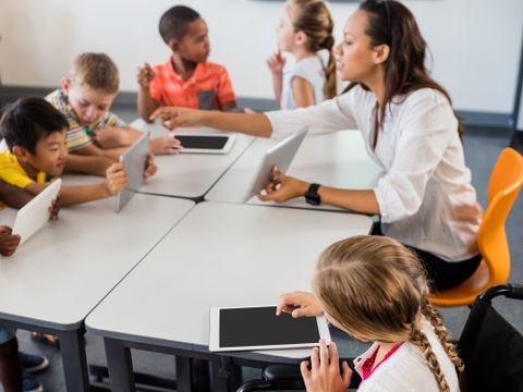 Eine Gruppe von Kindern sitzen mit Tablets in der Hand in einem Klassenraum, ihre Lehrerin zeigt etwas auf ihrem Tablet