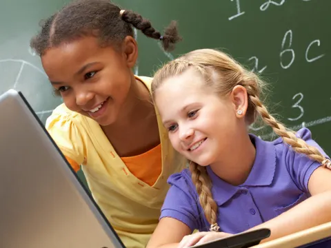 zwei Kinder vor einem Laptop mit Tafel im Hintergrund
