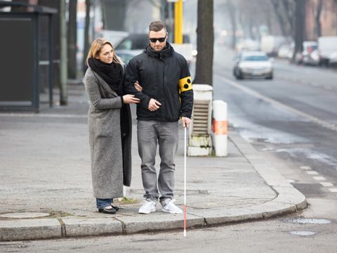 Eine junge Frau führt einen jungen Mann mit einem weißen Stock über die Straße