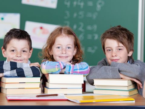 drei Kinder vor einer Tafel lehnen sich auf einen Stapel Bücher