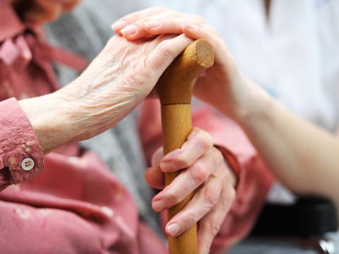 Eine alte Frau hält sitzend einen Gehstock mit der linken Hand umfasst und hat die rechte Hand auf den Griff gelegt. Darüber ist die Hand einer jüngeren Pflegeperson zu sehen.
