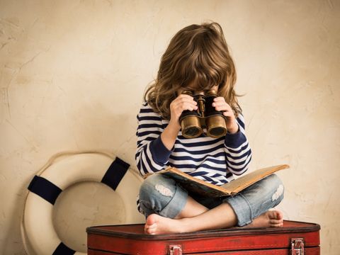 Kind sitzt auf einem roten Koffer und schaut durch ein Fernglas in ein Buch