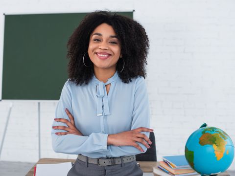 Junge Lehrerin lehnt lächelnd an ihrem Schreibtisch