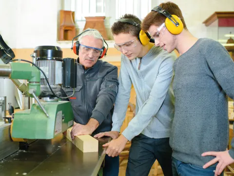 Zwei Auszubildende in einem Handwerksbetrieb, daneben ihr Ausbilder mit einem Stück Holz zum Zusägen. Alle tragen Ohrenschützer.