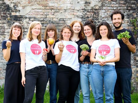 Bild zeigt das aus 8 Personen bestehende Projektteam von Restlos Glücklich welche vor einer Mauer stehen