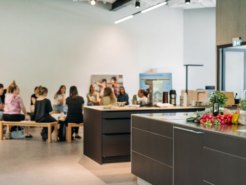 Ein großer Raum mit einer Küche im Vordergrund, im Hintergrund sitzen Menschen auf Bänken in einem Kreis