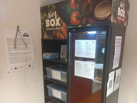 Kiez-Box im Stadtteilzentrum Zukunftshaus Wedding, ein Kühlschrank und ein Regal mit dem Aufdruck "Kiez Box" stehen in einer Zimmerecke