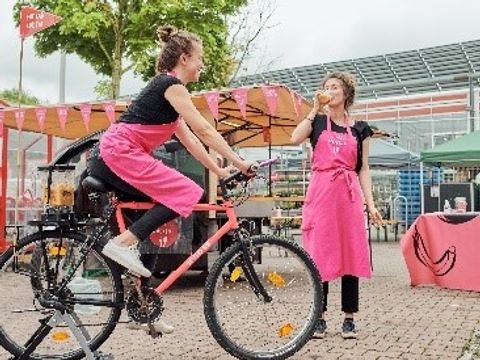 Eine Frau auf einem Smoothie-Bike, im Hintergrund trinkt eine Frau einen Smoothie