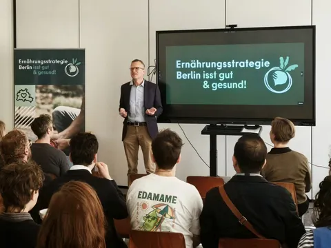 Ein Mann hält eine Rede, man sieht mehrere Zuschauerreihen von hinten, neben dem Redner steht ein Smartboard, auf welchem das Logo der Berliner Ernährungsstrategie zu sehen ist