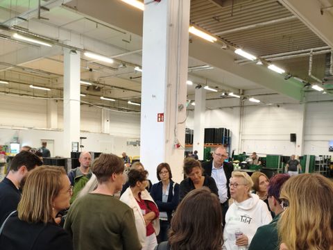 Eine Gruppe von Menschen steht in einer Halle im Kreis, eine Frau in der Mitte spricht zu der Gruppe