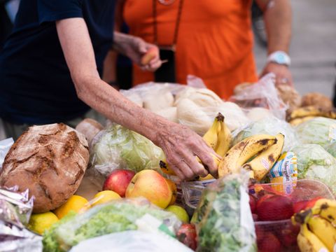 der Arm einer Person streckt sich über einen Tisch voller geretteter Lebensmittel, darunter Bananen, Äpfel und Brot