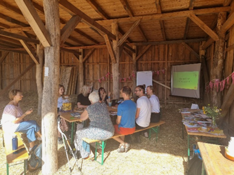 Eine Gruppe von erwachsenen Menschen sitzt in einem geräumigen offenen Holzschuppen und unterhält sich