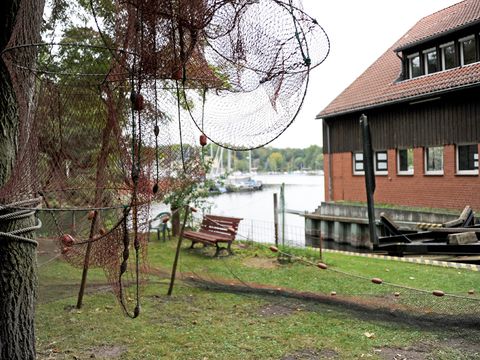 Reusen am Fischereiamt. Foto von Fischreusen, die zum Trocknen am Baum aufgehängt wurden. In Hintergrund sieht man das Dienstgebäude des Fischreriamtes und die Havel mit Segelbooten an einem Bootsanlegesteg