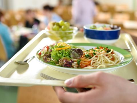 Person in einer Kantine hält ein Tablett mit Spaghetti und Tomatensauce, Salat und Weintrauben