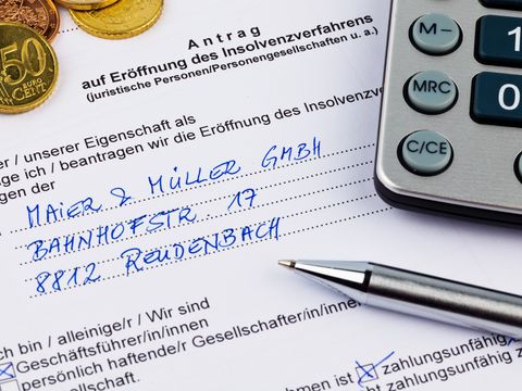 Ein ausgefüllter Antrag auf Eröffnung eines Insolvenzverfahrens. Am oberen Rand liegen mehrere Münzen. Auf der rechten Seiten ist ein Teil von einem Taschenrechner zu sehen. Direkt darunter liegt ein Kugelschreiber.