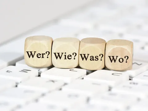 Auf einer Tastatur liegen vier Würfel mit den Aufschriften 'Wer?', 'Wie?', 'Was?', 'Wo?'