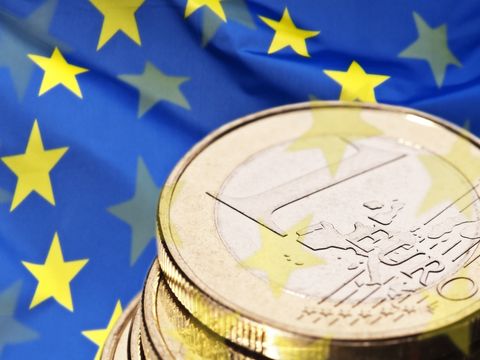 Übereinander liegende Ein-Euro-Münzen. Im Hintergrund sieht man die Europaflagge.