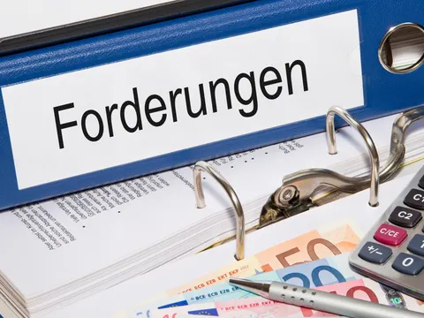 Auf einem geöffneten Aktenordner liegen ein Taschenrechner, ein Stift, mehrere Geldscheine und weitere Ordner mit der Aufschrift 'Forderungen'