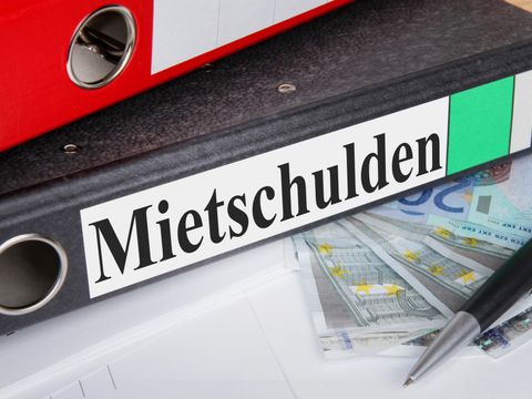 Ein Aktenordner mit der Aufschrift 'Mietschulden' liegt auf einem Schreibtisch. Auf ihm liegt ein weiterer Ordner, daneben ein Stift und mehrere Geldscheine.